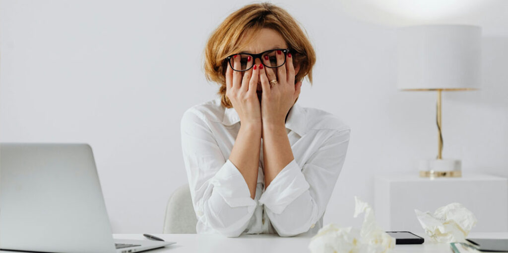 Femme au bureau, les mains sur les yeux, submergée par le stress et la surcharge de travail : l'importance d'une formation stress au travail